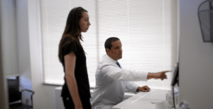 Male doctor in a white coat explains something on a computer screen to a standing female patient in a bright office setting.