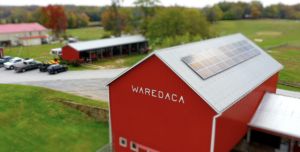 Aerial view of a large red barn with solar panels on its metal roof, surrounded by green fields, a driveway, and several parked vehicles nearby on a farm.